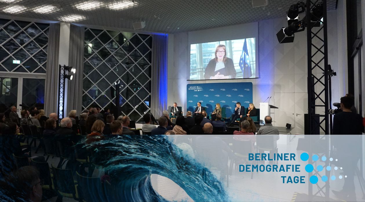 Audience in a futuristic room, an podium of 6, person speaking on a projected image. Logo of Berlin Demography Days