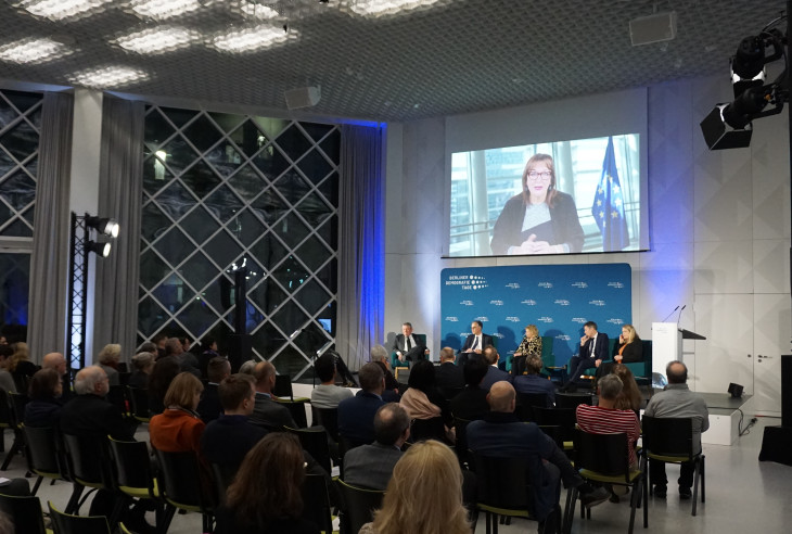 Podium discussion at Futurium Museum in Berlin. Rows of audience, a panel of 6, A person speaking on a projected image