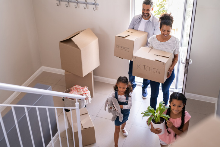 family with two children carrying boxes and plant in new home on moving day.