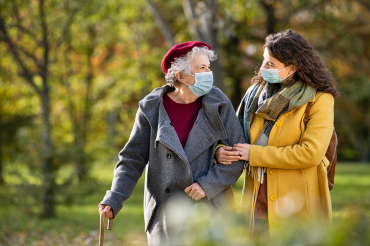 granddaughter walking with senior woman holding stick in park and wearing mask for safety against covid-19