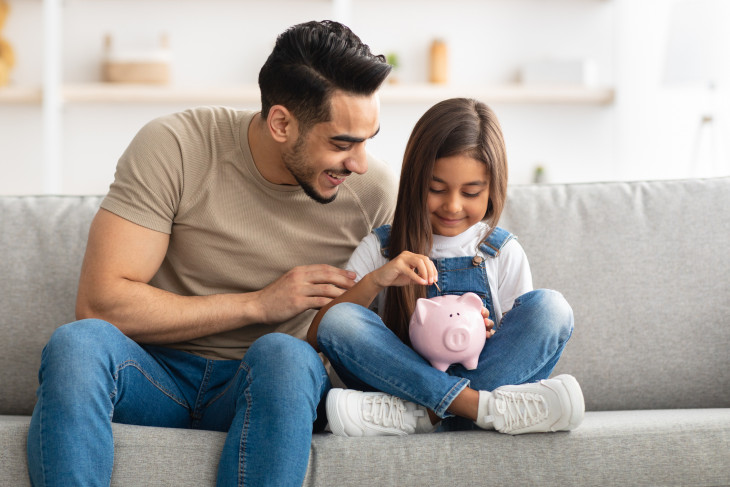 Portrait of cute smiling little girl putting coin in pink piggy bank, sitting with dad on the couch at home