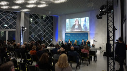 Podium discussion at Futurium Museum in Berlin. Rows of audience, a panel of 6, A person speaking on a projected image